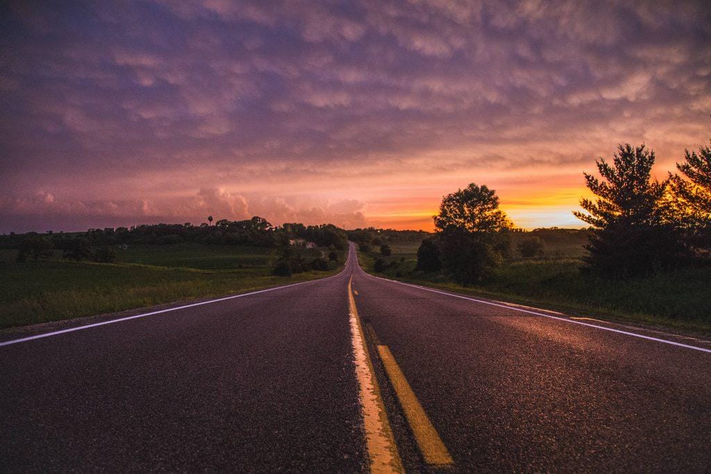 Long rural highway to the horizon under a purple-orange sunset, fields and trees — ideal backdrop for Hi‑Vac vacuum trucks.