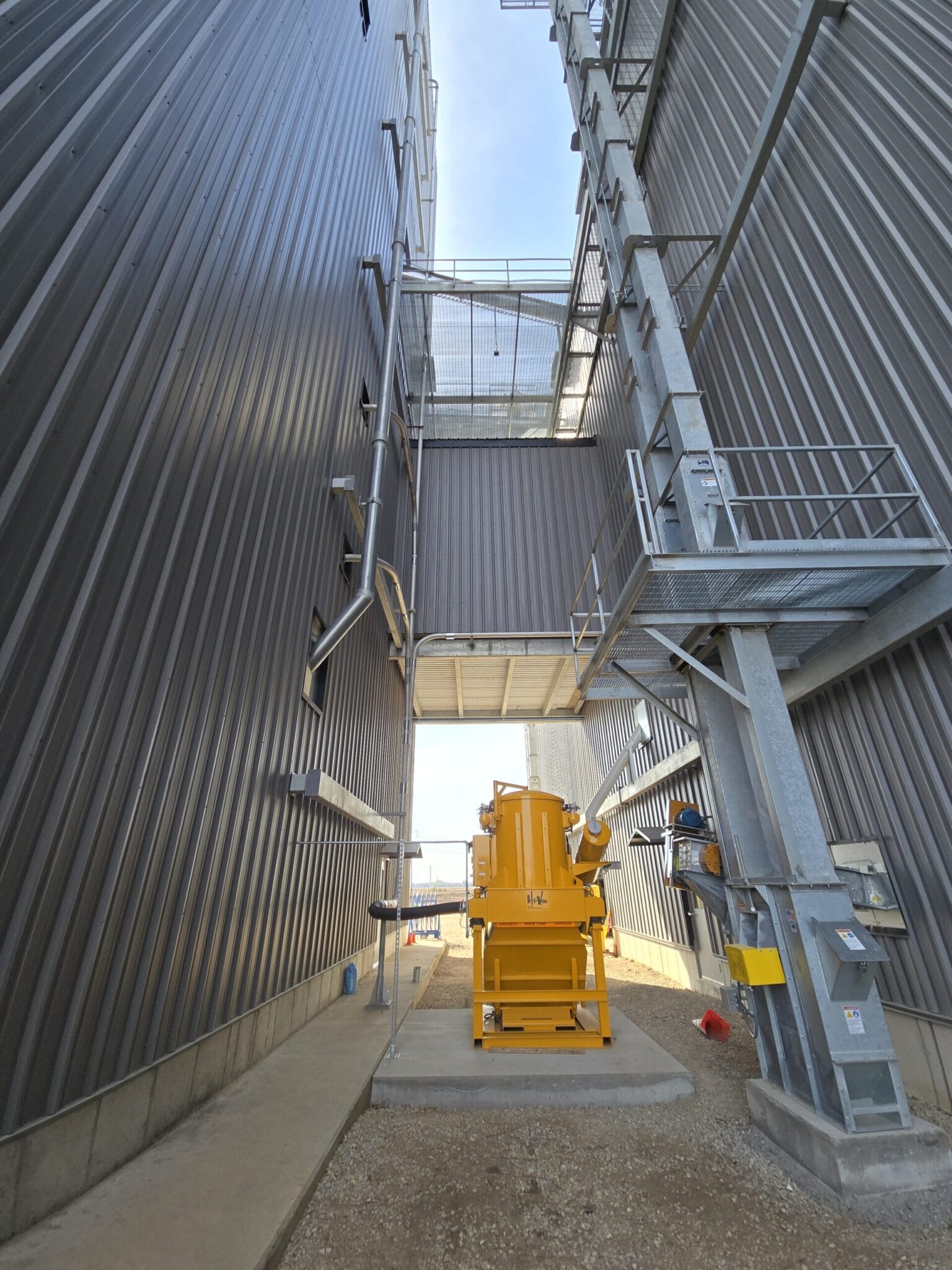 Yellow Hi‑Vac industrial vacuum unit centered between tall corrugated metal walls with overhead catwalk and steel ladder.