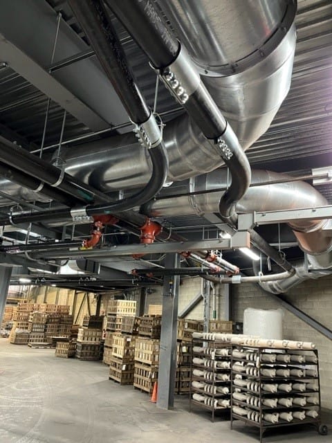 Overhead stainless ductwork and black vacuum hoses with red valves above warehouse racks of stacked ceramic molds, Hi-Vac