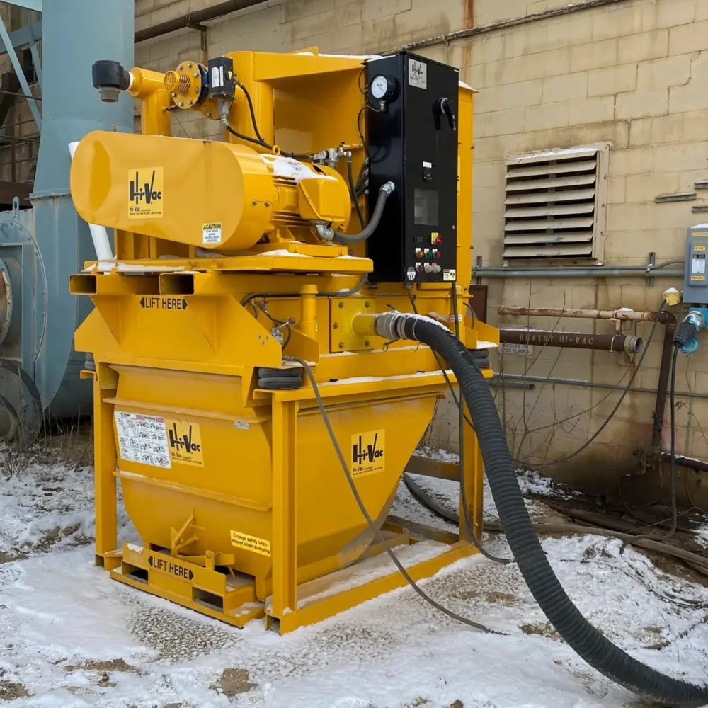 Yellow Hi‑Vac industrial vacuum hopper and motor skid with control panel and large black suction hose, dusted with snow