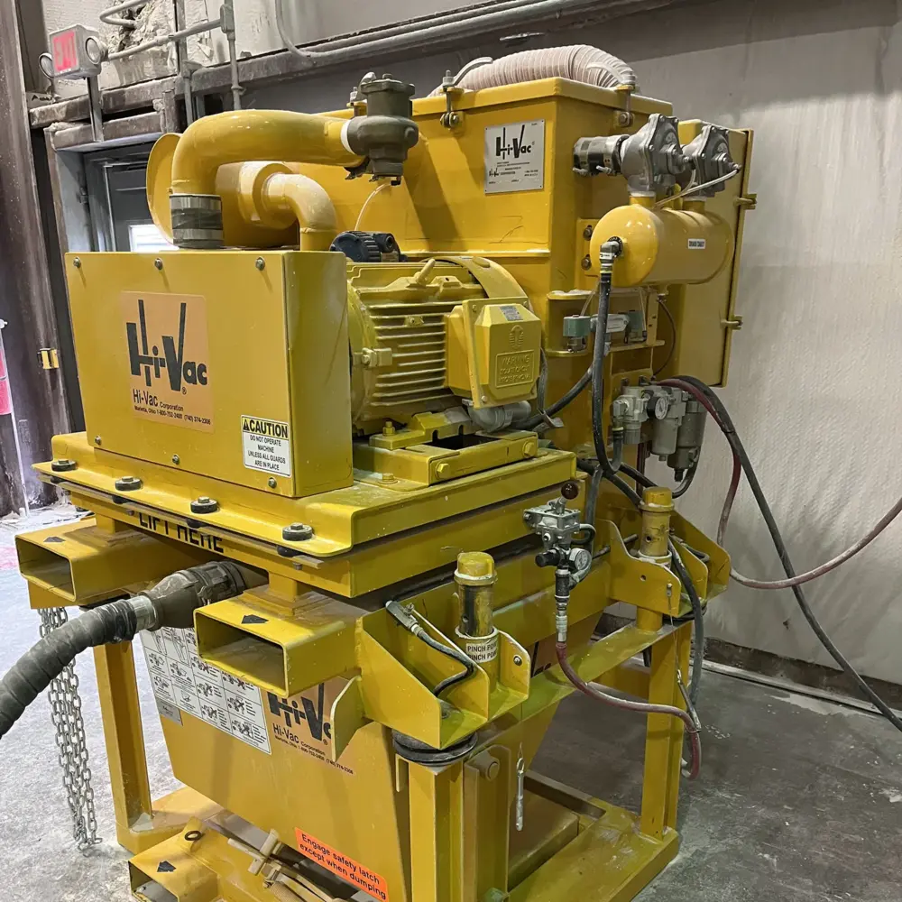 Yellow Hi-Vac industrial vacuum unit with electric motor, hoses and control valves mounted on a steel frame in a dusty shop